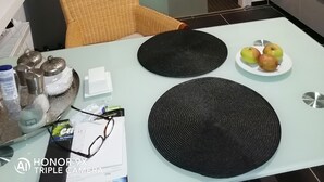 Une cuisine compacte comprenant un réfrigérateur, une table à manger avec un set de table en fibres tressées, une assiette de fruits et une chaise en osier.