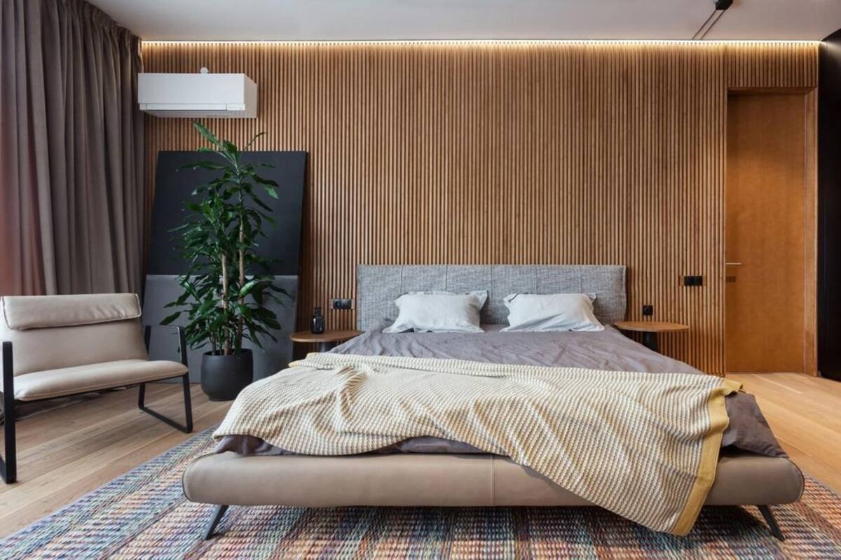 A modern bedroom with a large bed, a side table, a chair, a potted plant, and a wooden panel wall.