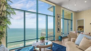 A spacious living room with a large window overlooking the ocean, a round coffee table with a plant, and a beige sofa. - Jr Penthouse 29th Floor Tallest Building SPI (South Padre Island)