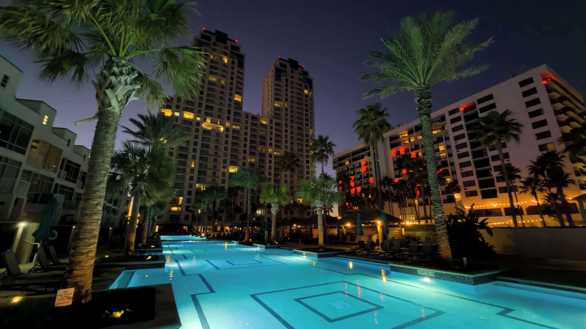 A swimming pool with a geometric design, surrounded by palm trees and modern buildings with illuminated windows.