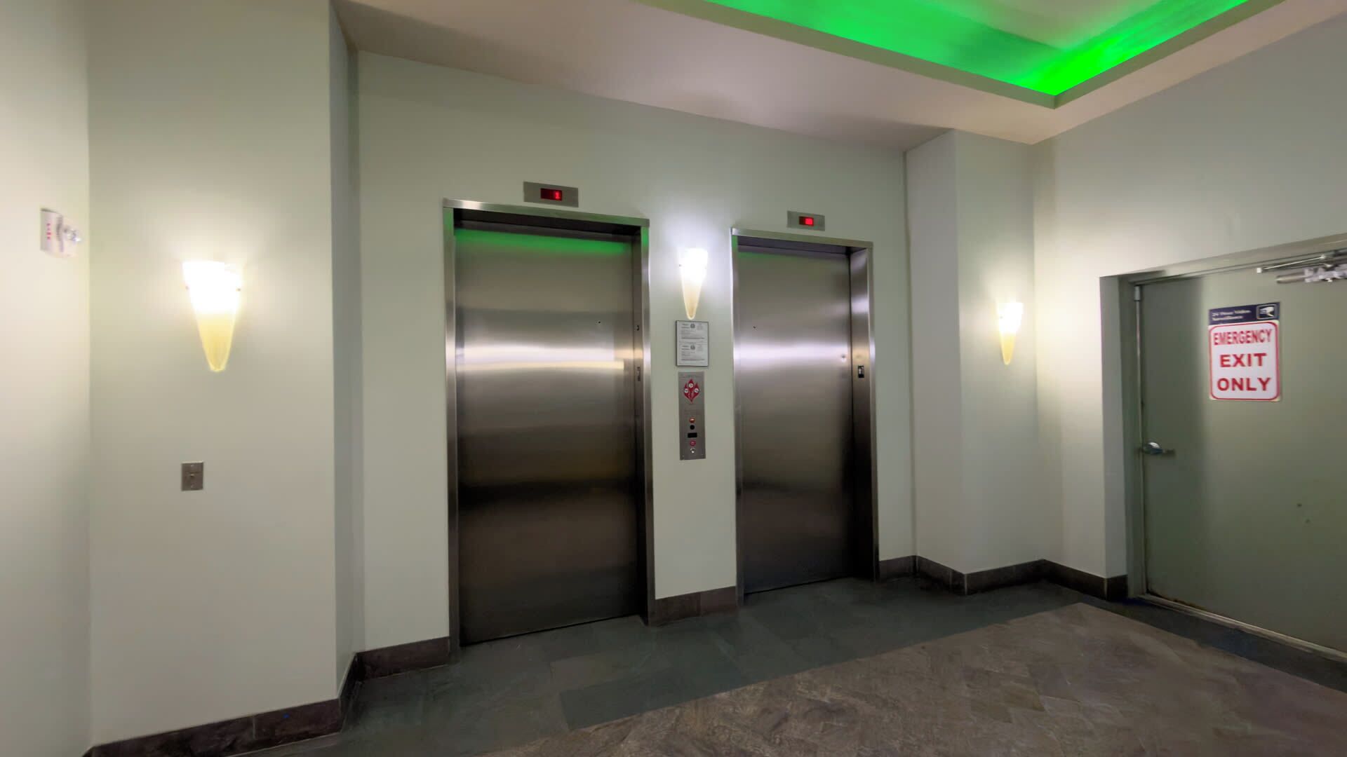 An elevator area with two elevators, a green exit sign, and a wall-mounted light fixture.