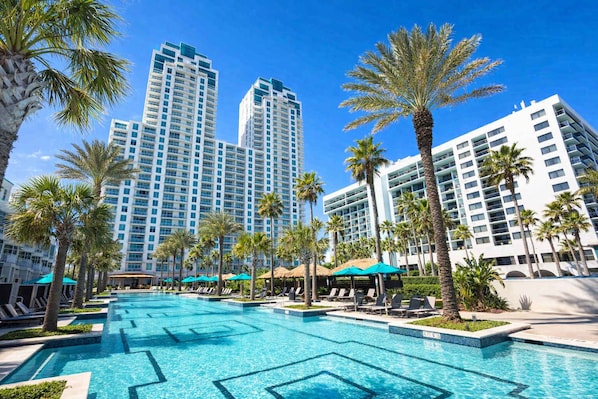 Un hotel con zona de piscina, palmeras y edificios altos al fondo.