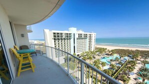 A balcony with a yellow chair and table, overlooking a beach and a resort with multiple swimming pools and palm trees.