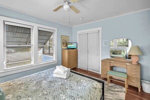 A bedroom with a bed, a dresser, a television, a desk, a chair, a ceiling fan, and a window with blinds.