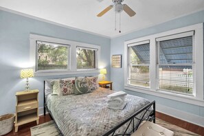 Un dormitorio con cama, dos mesitas de noche, ventilador de techo y ventanas amplias con persianas.