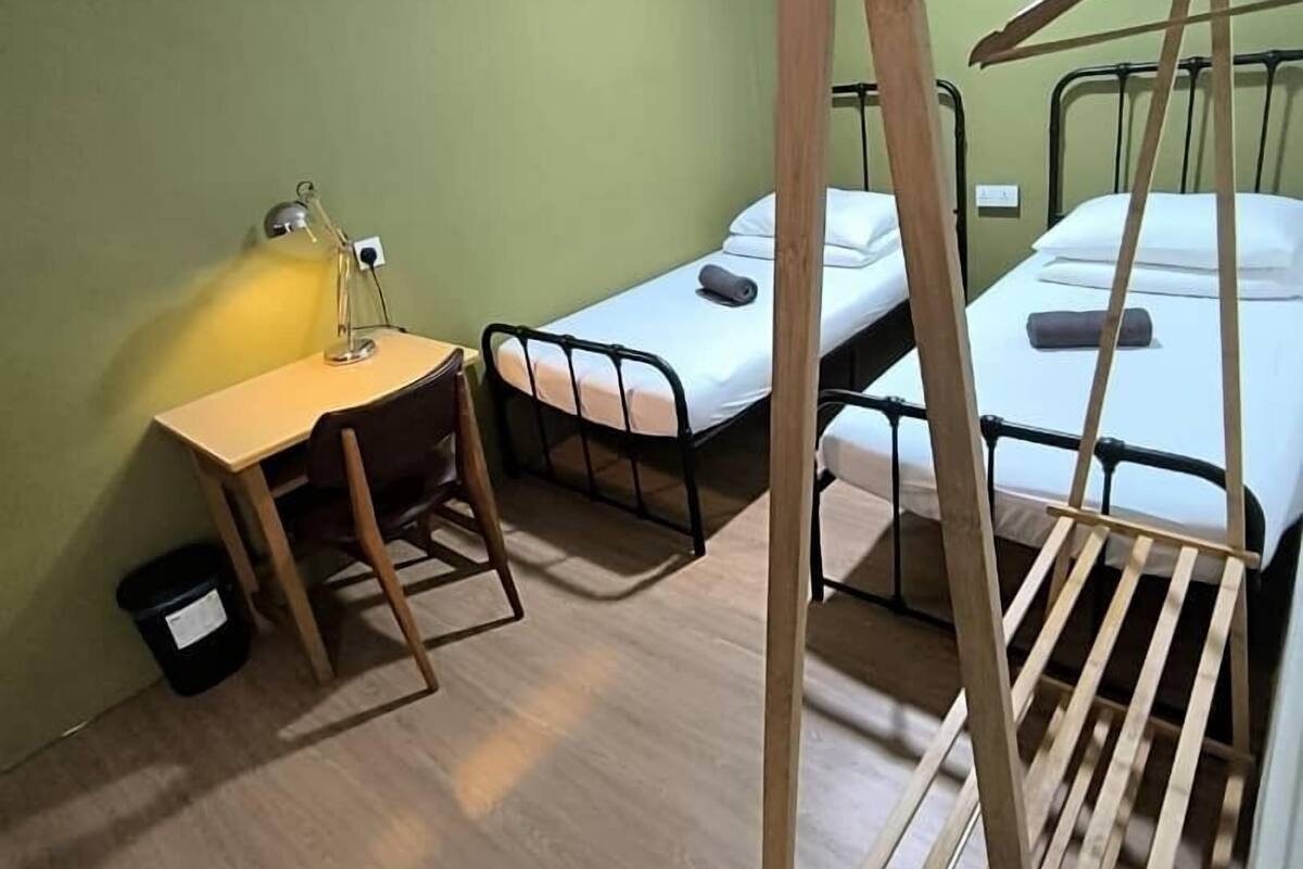 Una habitació de dormitori amb dos llits individuals, un escriptori de fusta amb una cadira, una prestatgeria per a la roba i un petit cubell de brossa.