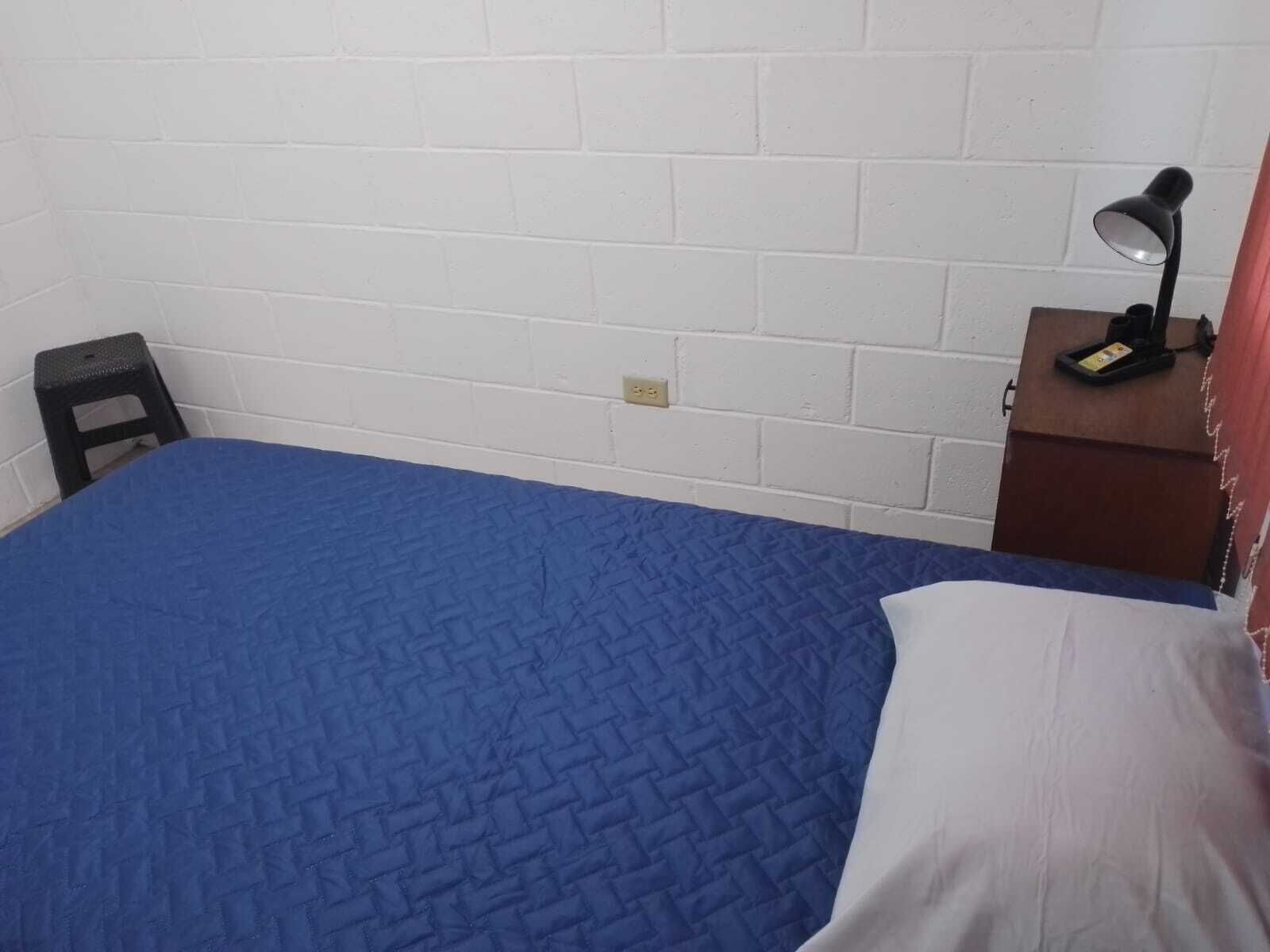 Ein kleines Zimmer mit einem Einzelbett, einer blauen Bettdecke, einem schwarzen Hocker, einem Schreibtisch mit Lampe und einer weißen Wand.