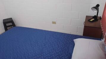 Ein kleines Zimmer mit einem Einzelbett, einer blauen Bettdecke, einem schwarzen Hocker, einem Schreibtisch mit Lampe und einer weißen Wand.