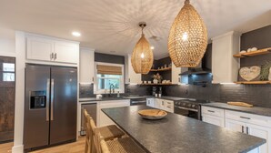 A modern kitchen with a stainless steel refrigerator, a large island with a bowl, wicker chairs, and hanging pendant lights. - Little Canal, Pool, Hot Tub, Cold Plunge, Sauna (Santa Rosa Beach)