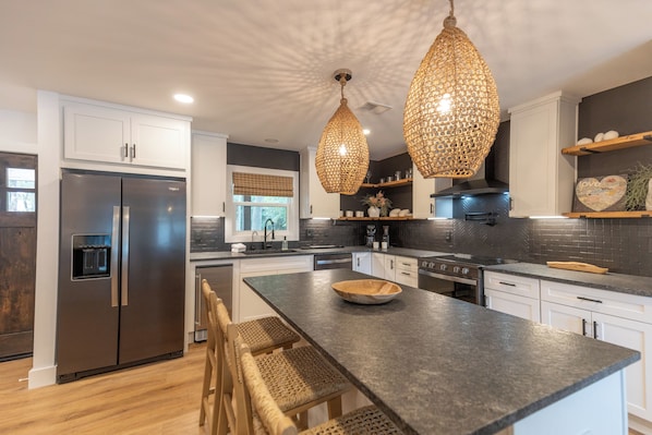 A modern kitchen with a stainless steel refrigerator, a large island with a bowl, wicker chairs, and hanging pendant lights.