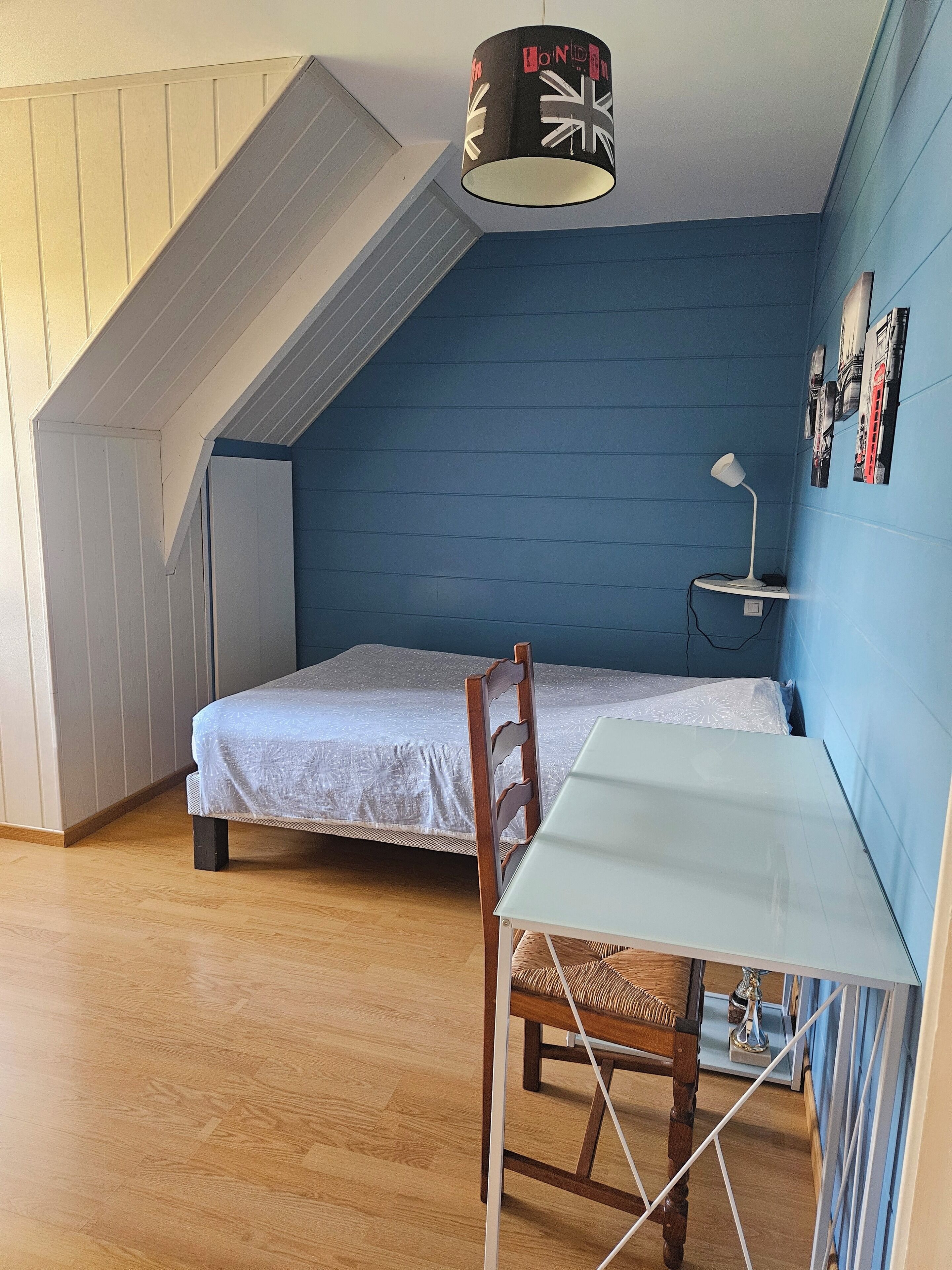 Een kleine, compacte kamer met een schuine kap, een enkel bed, een bureau met stoel en een lamp.