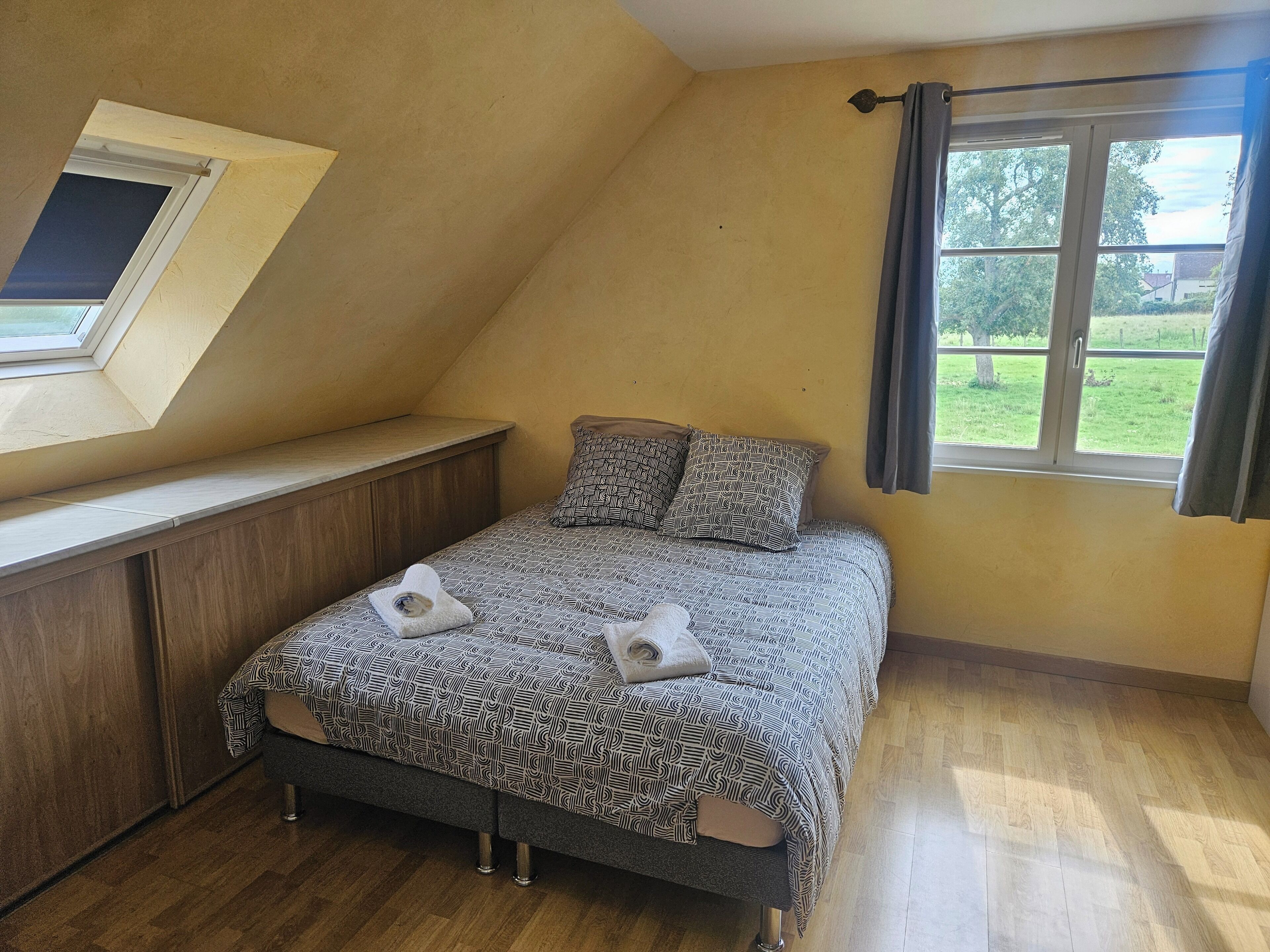 A bedroom with a sloped ceiling, a bed with patterned bedding, a window with curtains, and a view of greenery outside.