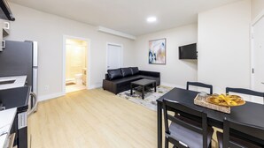 Uma sala de estar moderna com um sofá preto, uma mesa de jantar com cadeiras, uma televisão de tela plana na parede e uma área de cozinha com geladeira e micro-ondas.