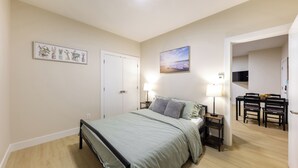 Quarto com cama, mesas de cabeceira, escrivaninha com cadeiras, um quadro e uma porta.
