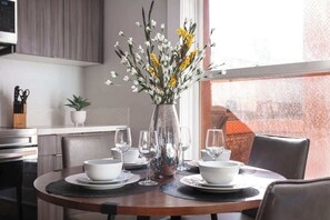 A dining area with a round table set for two, a vase with flowers, and a view of the city through a large window. - The Penn Hotel -  Vibrant Gaslamp Suite Convenient Central Location (San Diego)