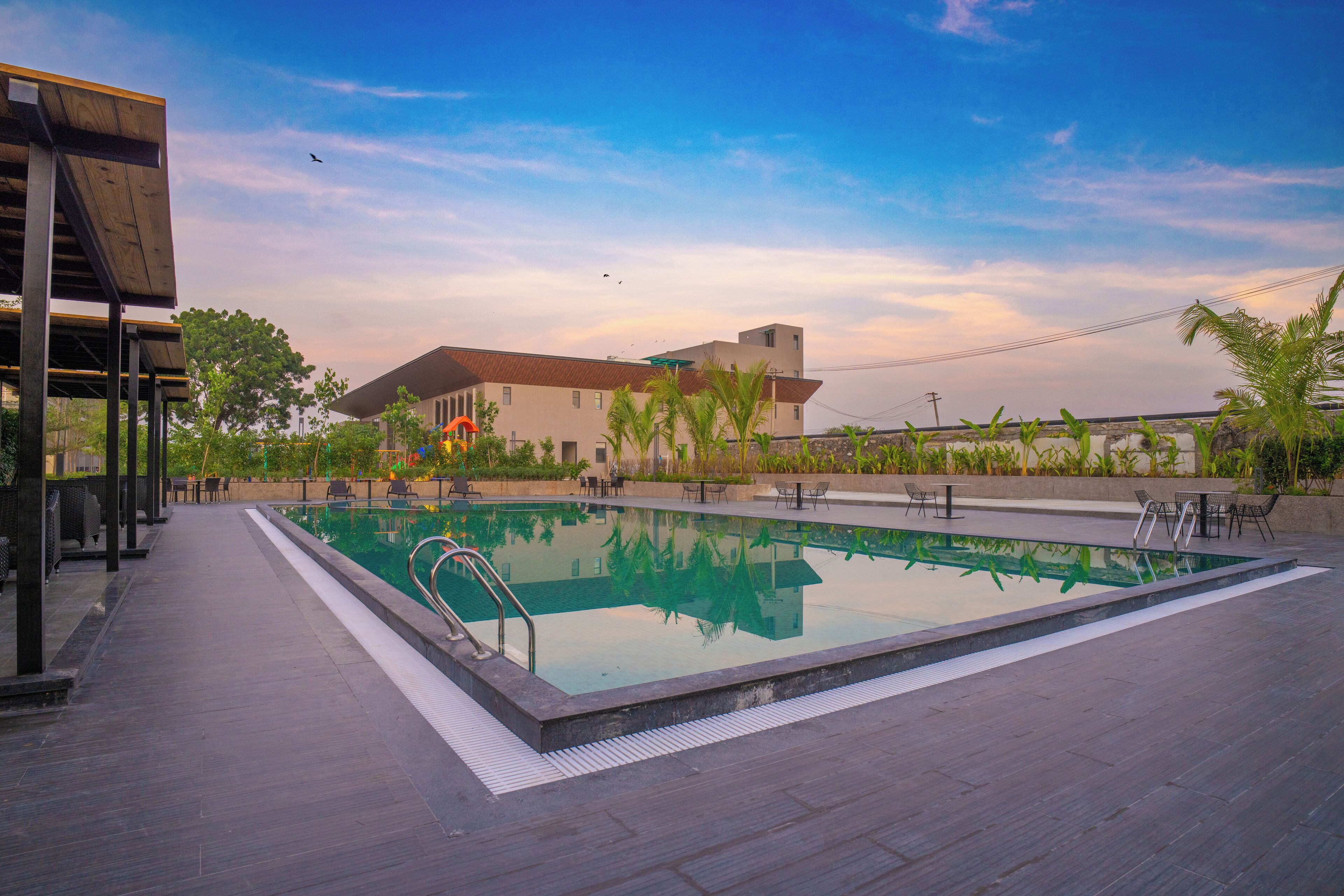 A swimming pool with a wooden deck, outdoor seating, and a building in the background.