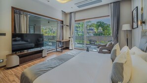 A modern hotel room with a large bed, a flat-screen TV, a balcony with a view of greenery, and a seating area with a chair and a table. - AMARTARA THE RESORT (Abu Road)