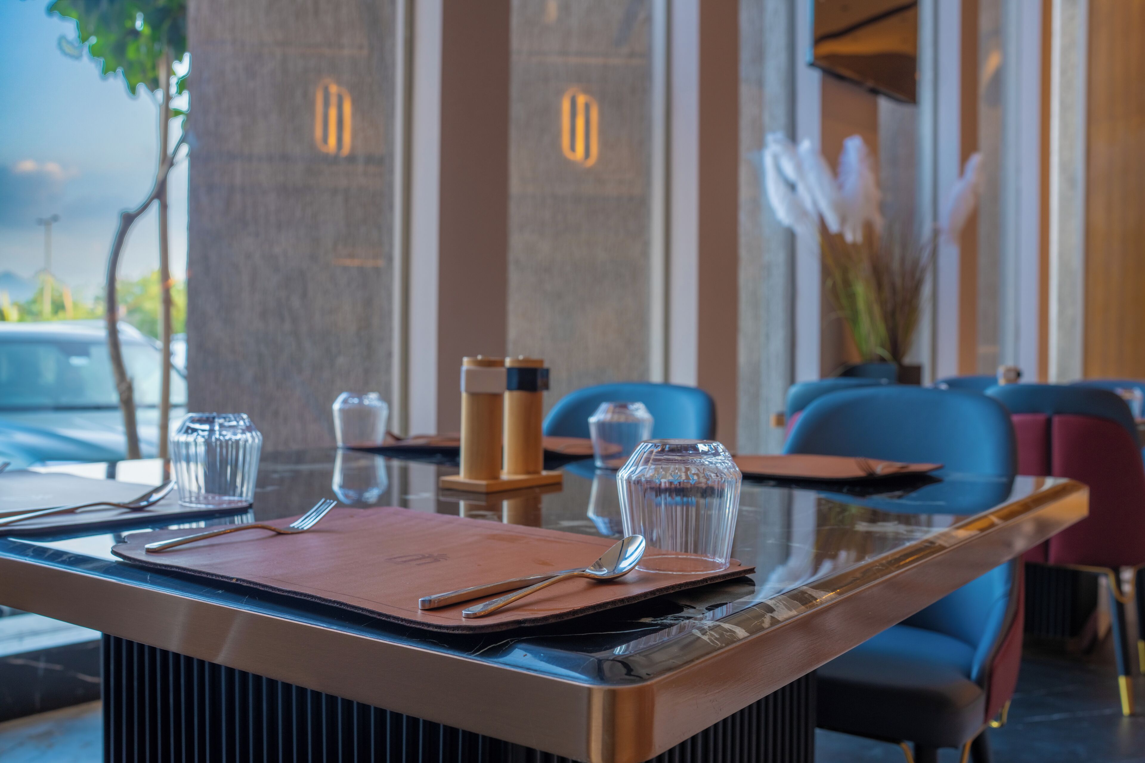 A restaurant setting with a table set for two, featuring a clear glass top, a brown menu, and cutlery.