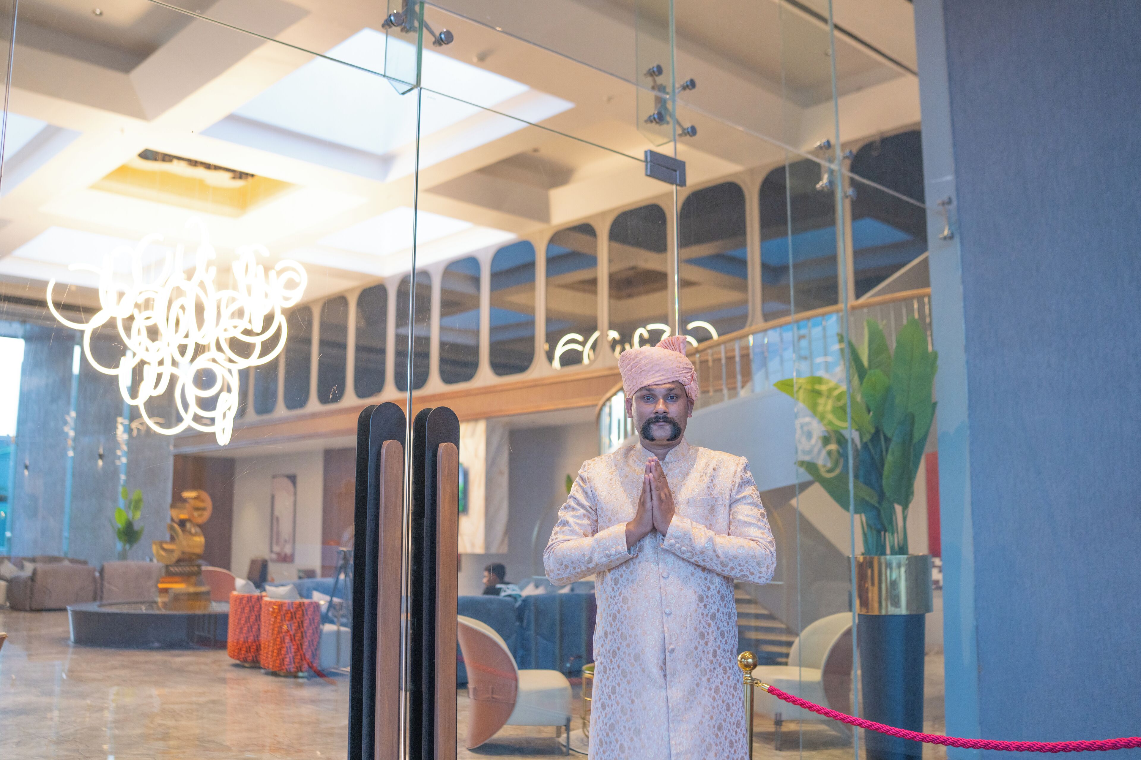 Ein Mann in traditioneller Kleidung steht in einer modernen Lobby mit Glaswänden, einem Chandelier und einer Treppe im Hintergrund.