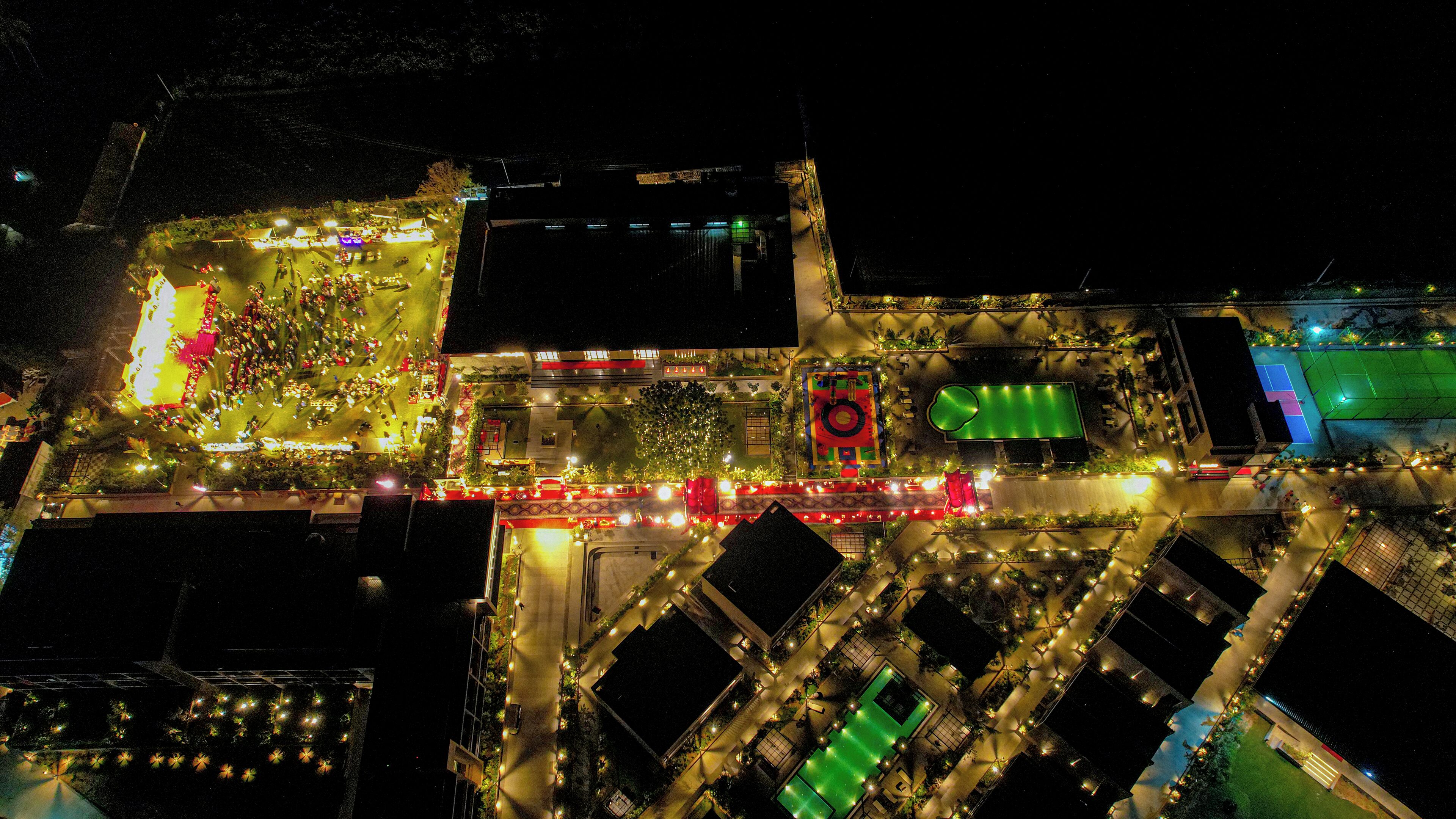 Une vue aérienne d’un espace extérieur bien éclairé, comprenant un court de tennis, des espaces de détente et un bâtiment central doté d’un élément circulaire rouge.