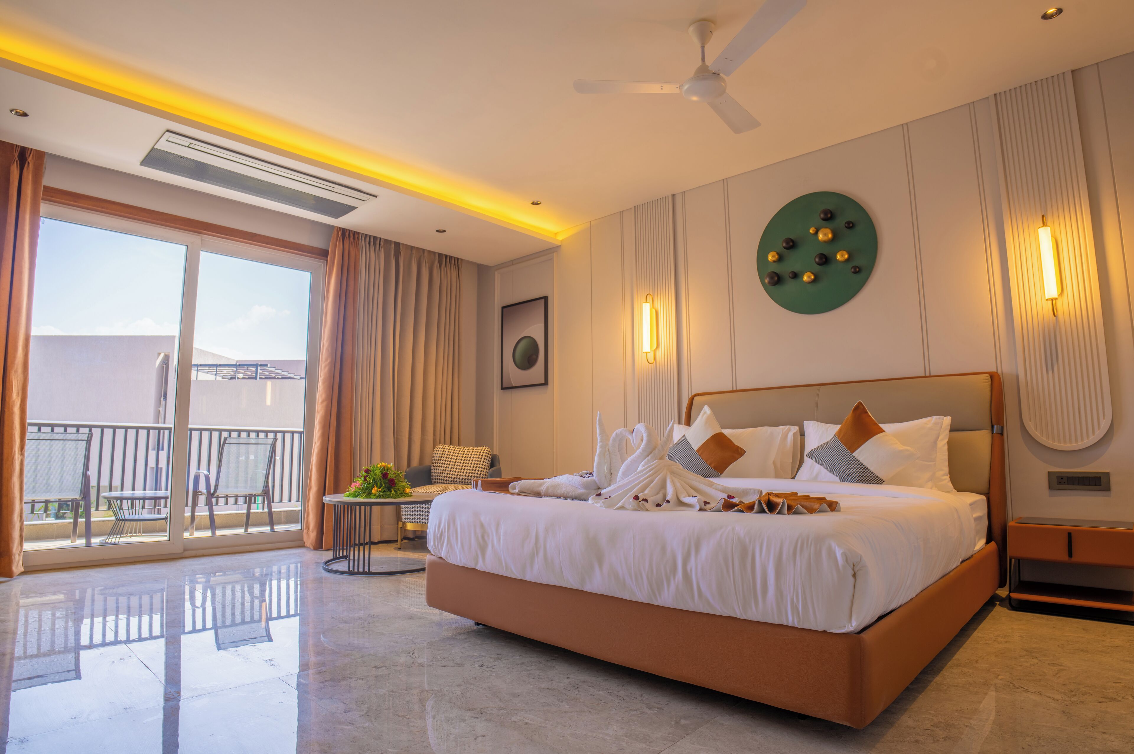 A spacious hotel room with a large bed, a balcony with chairs, and a ceiling fan.