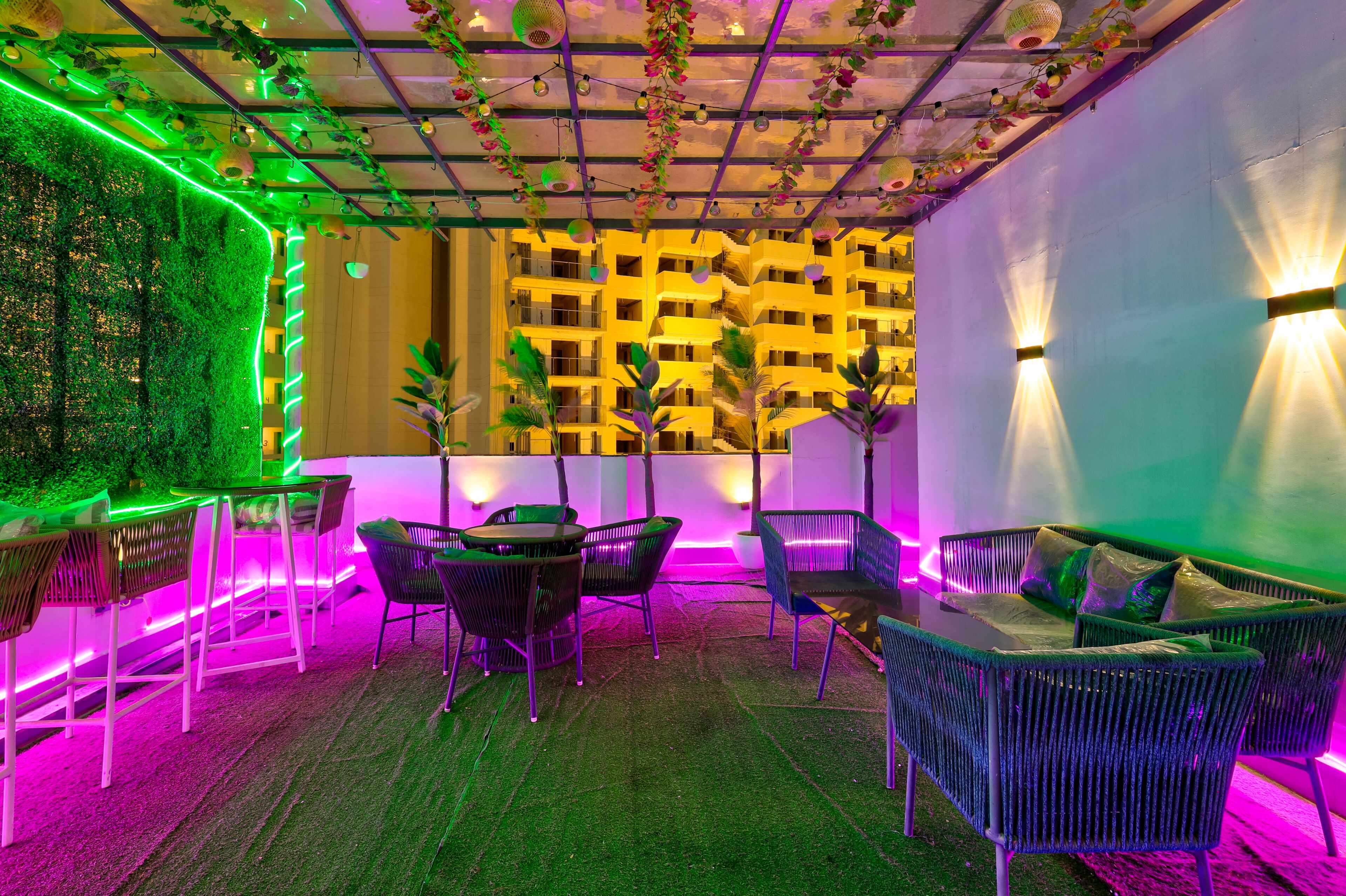 A modern outdoor seating area with green and purple ambient lighting, a patterned wall, and a ceiling with hanging lights.