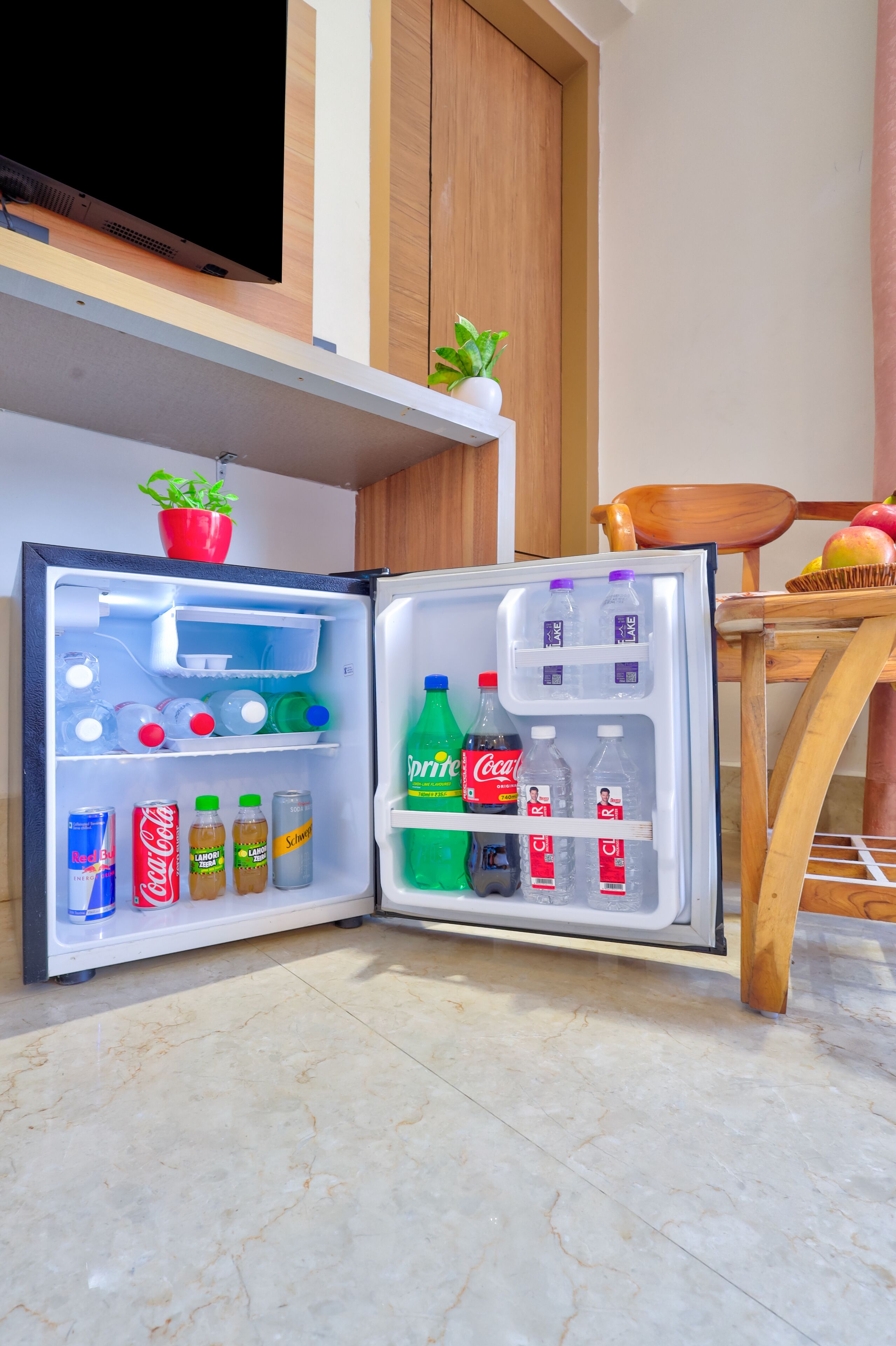 An open refrigerator stocked with beverages and snacks, a potted plant on the countertop, and a wooden chair with fruits.