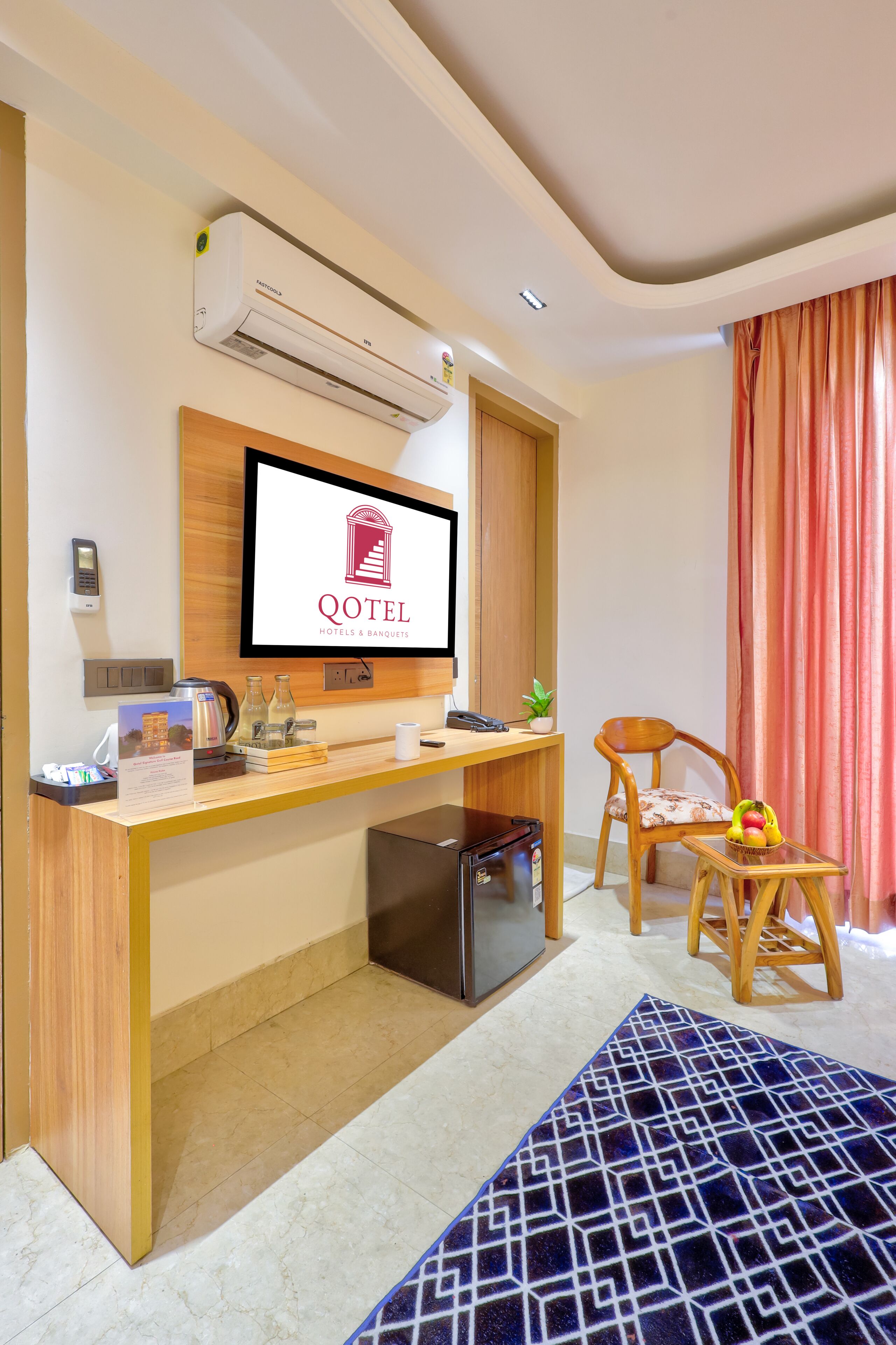 A hotel room with a desk, a television displaying the hotel logo, a refrigerator, a chair, and a window with curtains.