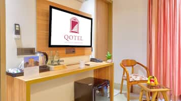 A hotel room with a desk, a television displaying the hotel logo, a refrigerator, a chair, and a window with curtains.