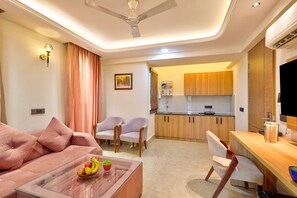A modern living room with a sofa, chairs, a glass coffee table with fruit, and a kitchen area with wooden cabinets and a dining table.