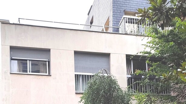 A multi-story building with a flat facade, a visible entrance, and a small garden area with pink flowers.