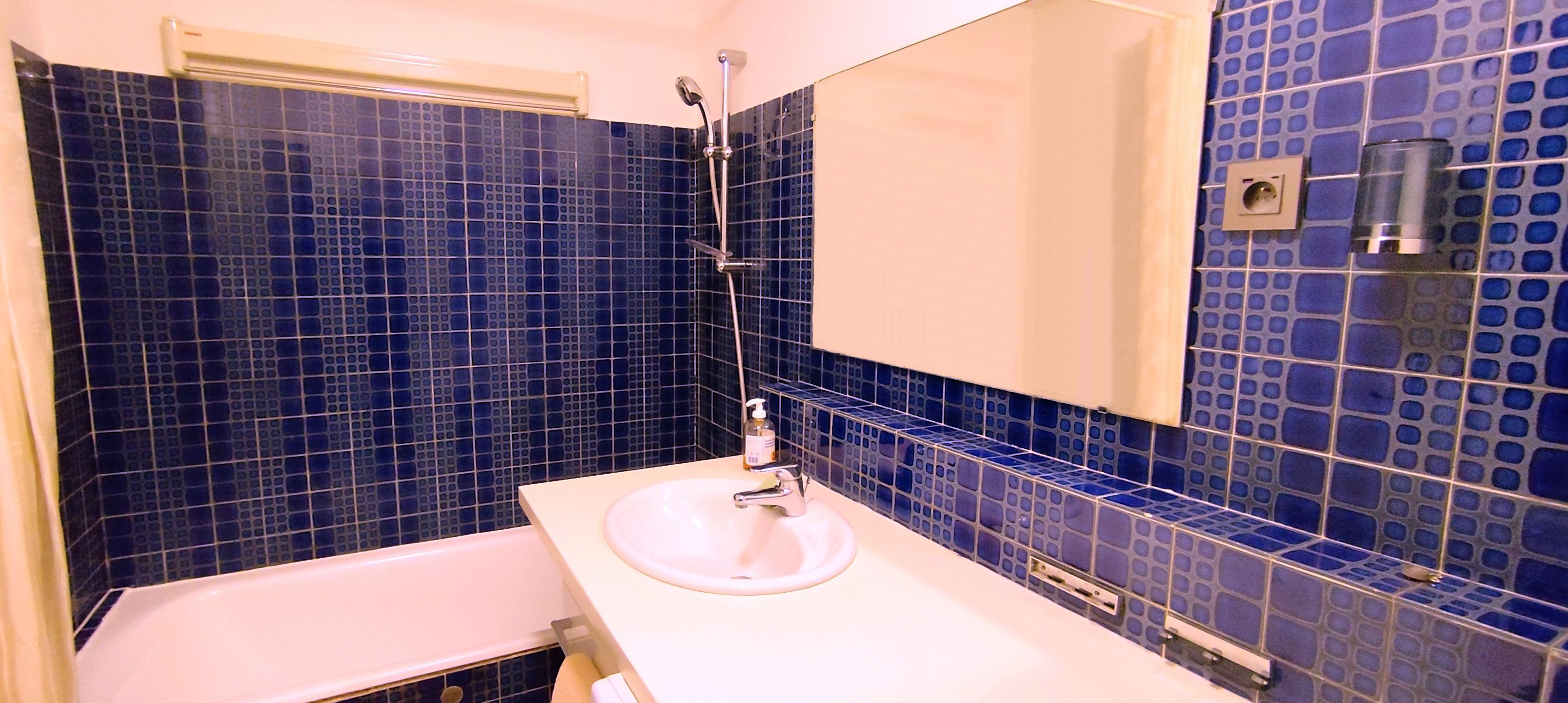 Une salle de bain avec un lavabo blanc, un miroir et une douche. Les murs sont recouverts de carreaux de mosaïque bleus.