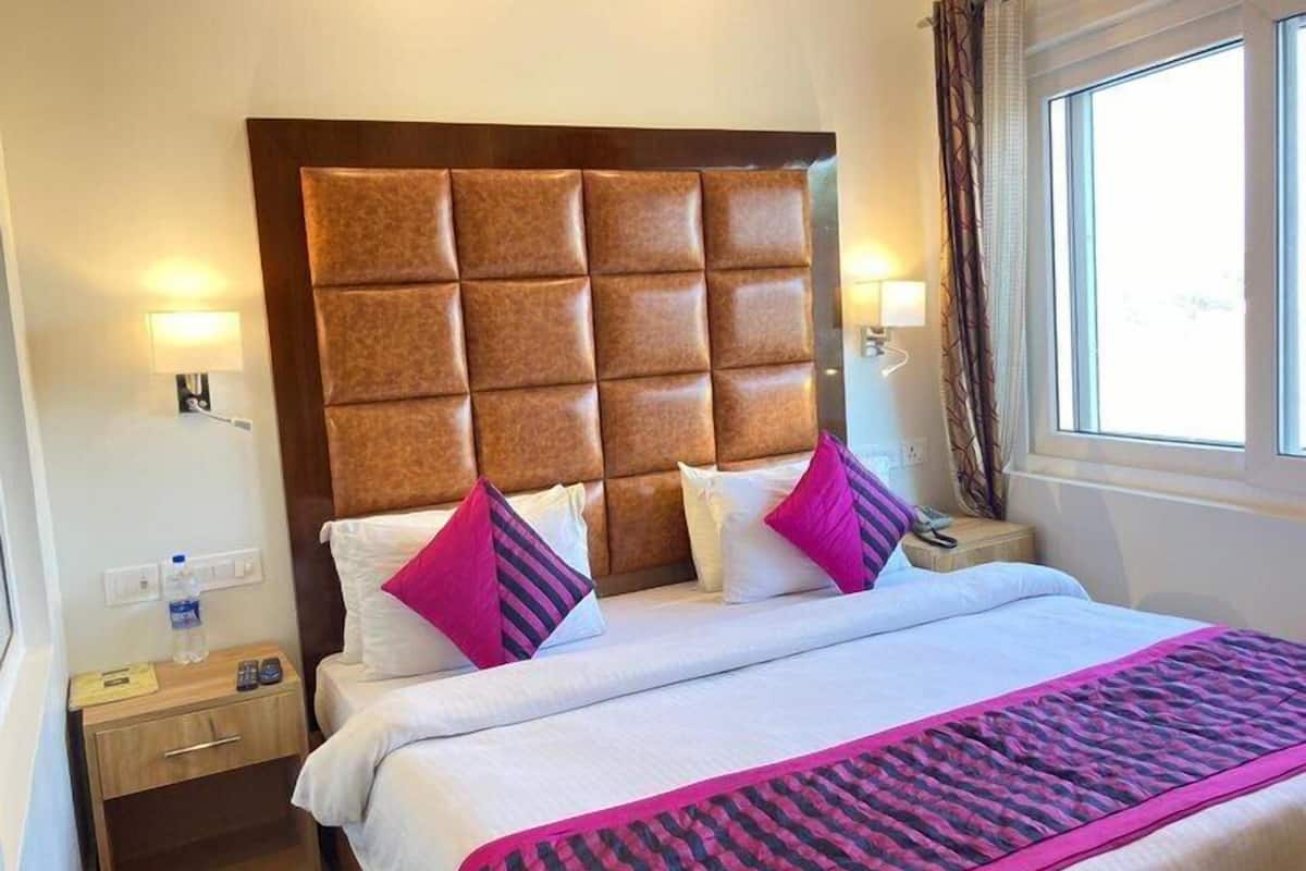 Ein Hotelzimmer mit einer gesteppten Kopfteil, einem Bett mit weißen Leinen und rosa Akzenten, einem Nachttisch aus Holz mit einer Lampe und einem Fenster mit Vorhängen.