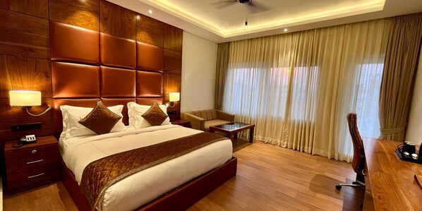 A hotel room with a large bed, wooden headboard, two bedside tables with lamps, a small desk with a chair, and a large window with curtains. - Hotel Guru Surbhi (Paonta Sahib)