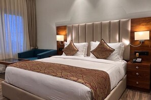 A hotel room with a large bed, two bedside tables with lamps, a blue armchair, and a view of the outdoors through the window. - Hotel Guru Surbhi (Paonta Sahib)