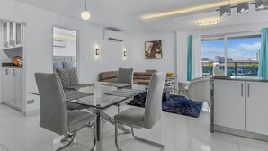 Een moderne woonkamer met een glazen eettafel, grijze stoelen, een bruine bank en uitzicht op de stad door grote ramen.