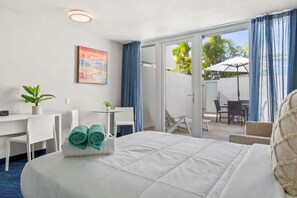 Ein Hotelzimmer mit Bett, Schreibtisch, Stuhl, einem kleinen Tisch, einer Topfpflanze und Blick auf den Außenbereich mit Terrasse und Sonnenschirm.
