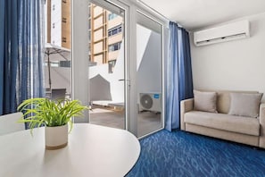 Habitación de hotel con sofá, una mesa de comedor con planta en maceta, puerta corrediza de vidrio a un espacio exterior y unidad de aire acondicionado en la pared.