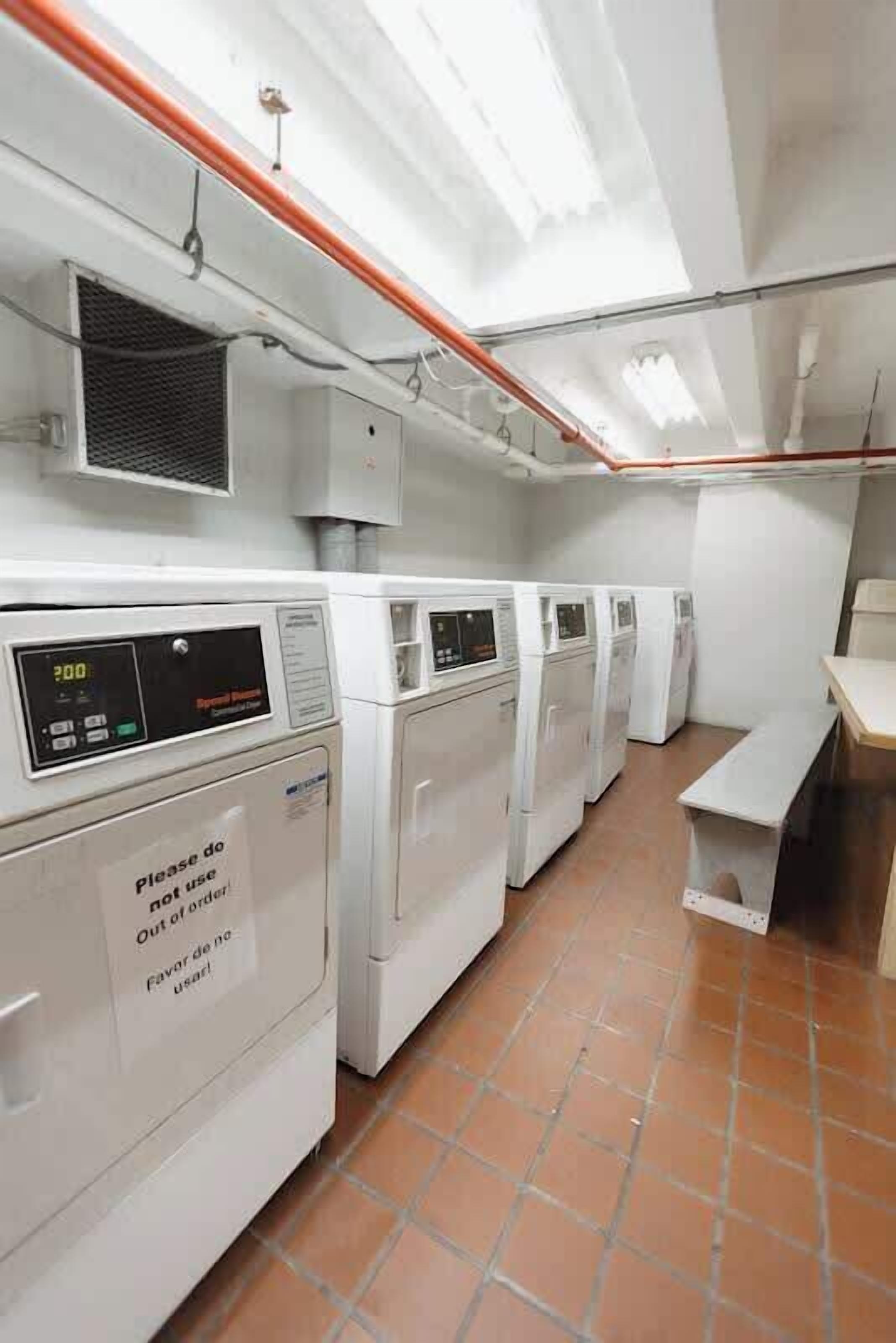 Une laverie équipée de plusieurs machines à laver alignées contre le mur, d’un panneau de commande doté d’un écran numérique et d’une affiche indiquant qu’il ne faut pas utiliser les machines.
