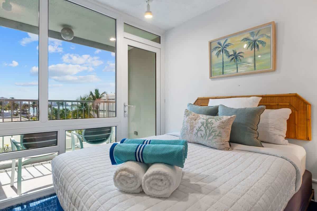 A bedroom with a large window, a bed with rolled towels, and a painting of palm trees.