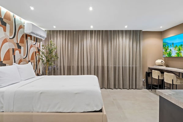 A modern hotel room with a large bed, a desk with two chairs, an air conditioning unit, and a large window with curtains.