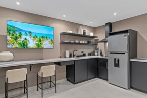 A modern kitchen with a large island, bar stools, a refrigerator, and a wall-mounted TV displaying a tropical beach scene.