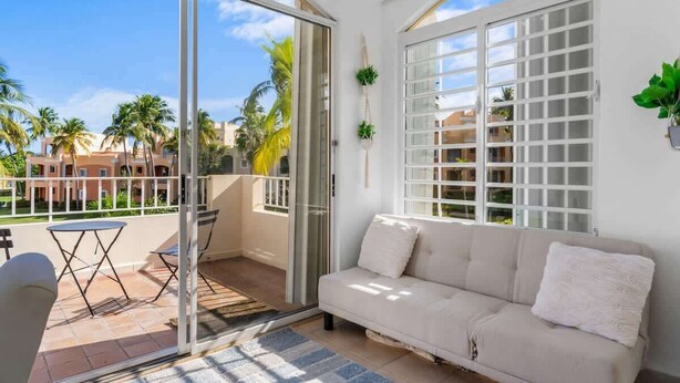 A balcony with a sofa, a table, and chairs, overlooking a residential area with palm trees and houses. - Studio w/ King Bed & Balcony (Palmas del Mar)
