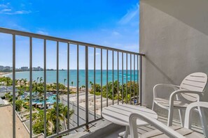 A balcony with a view of a beach and pool area, white chairs, and a railing. - Beachfront Studio | 2 Queen Beds + Parking (Carolina)