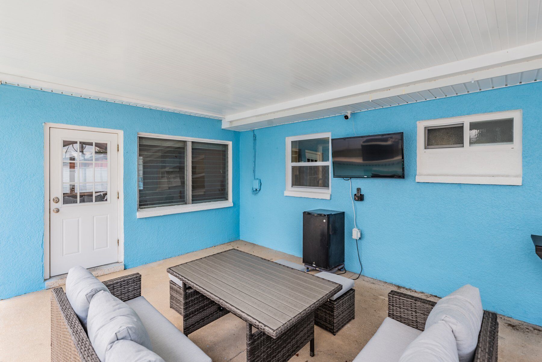 Eine überdachte Terrasse mit blauer Wand, weißer Tür, Tisch und Korbmöbeln.