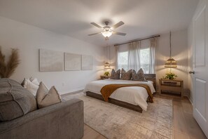 Une chambre à coucher avec un lit, un ventilateur de plafond, une table de chevet, une lampe et une fenêtre avec des rideaux.