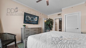 A bedroom with a bed, a ceiling fan, a TV mounted on the wall, a dresser, a bench, and a potted plant. - Ocean Club 406 | Enjoy Views of the Gulf! (Biloxi)