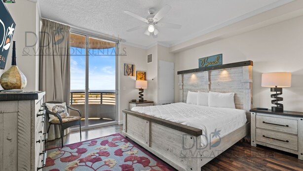 A bedroom with a bed, a nightstand, a lamp, a chair, a window with a view, and a ceiling fan. - Ocean Club 406 | Enjoy Views of the Gulf! (Biloxi)