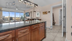 A modern kitchen with granite countertops, wooden cabinets, and a built-in island. There is a dining area with a table and chairs, and a view of the city through large windows. - Ocean Club 406 | Enjoy Views of the Gulf! (Biloxi)