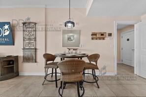 Een eetruimte met een ronde tafel, vier rieten stoelen, een hanglamp en een wanddecoratie.
